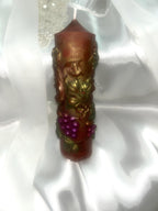 May include: A brown pillar candle with gold accents and a grape vine design. The candle features a cluster of purple grapes and green leaves. The candle is cylindrical and has a white wick.