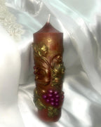 May include: A decorative pillar candle with a brown base and a gold-colored top. The candle is adorned with gold-colored leaves and a cluster of purple grapes. The candle is approximately 6 inches tall.