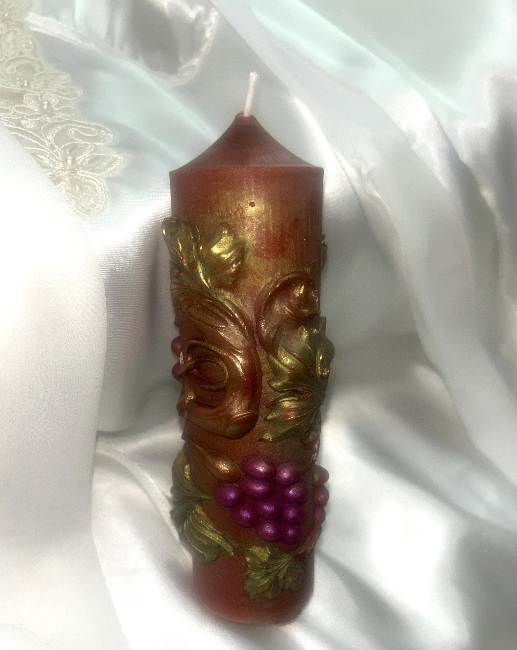 May include: A decorative pillar candle with a brown base and a gold-colored top. The candle is adorned with gold-colored leaves and a cluster of purple grapes. The candle is approximately 6 inches tall.