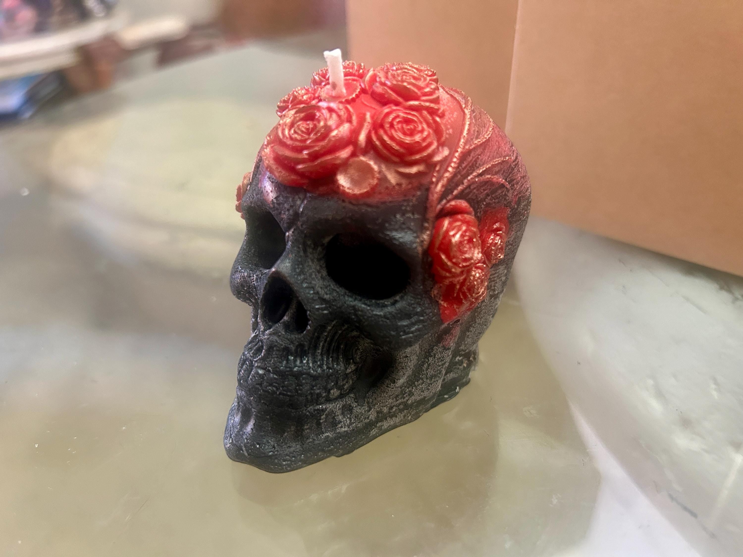 May include: A black skull-shaped candle with red rose accents on the forehead and sides. The candle has a white wick and is set against a neutral background. The skull's details are finely sculpted.